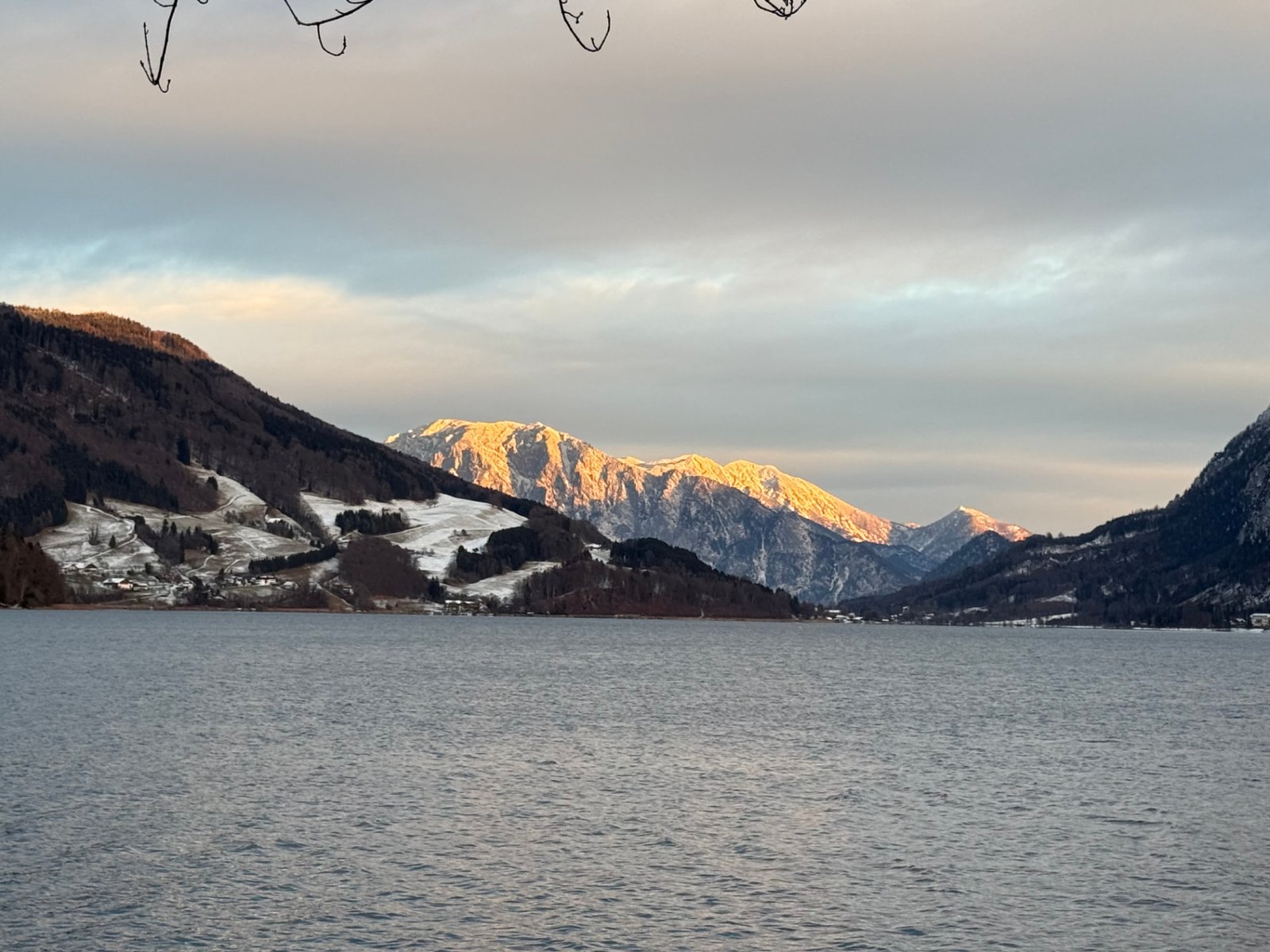 Sunlit mountain range beyond a lake beneath a cloudy sky.